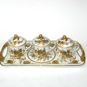 May include: A white porcelain tea set with gold floral designs. The set includes three lidded cups with handles, a serving tray, and gold accents. The tray has handles and a floral pattern.