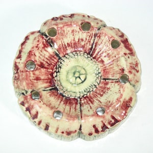 May include: A ceramic flower-shaped dish with a red and white glaze. The dish has eight holes around the center for holding flowers.
