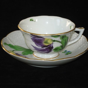 May include: A white porcelain teacup and saucer set with a floral design. The cup and saucer feature a purple and yellow flower with green leaves and a gold trim.