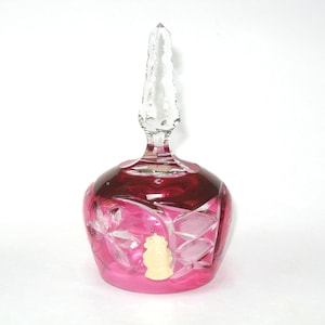 May include: A pink glass bell with a clear crystal top. The bell has a cut glass design and a small label on the side.