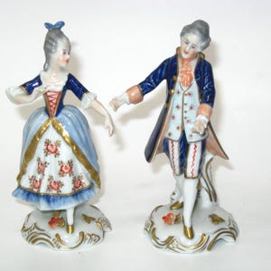 May include: Two porcelain figurines depicting a pair in 18th-century attire. The figure on the left wears a blue and white dress with floral patterns and gold trim. The figure on the right wears a blue coat, white vest with floral details, and white breeches.