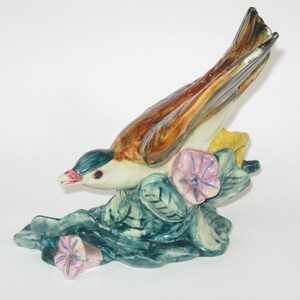 May include: A ceramic figurine of a brown and white bird with green and yellow accents perched on a bed of green leaves and pink flowers.