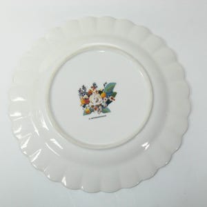 May include: White ceramic plate with a scalloped edge. The center of the plate features a floral design with a variety of flowers in shades of orange, yellow, blue, and white. The plate is likely for serving food.