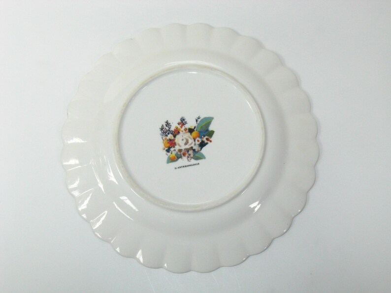 May include: White ceramic plate with a scalloped edge. The center of the plate features a floral design with a variety of flowers in shades of orange, yellow, blue, and white. The plate is likely for serving food.