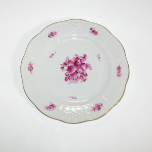 May include: White porcelain plate with a scalloped edge and gold trim. The plate features a central floral design and smaller floral accents in shades of pink. The plate has a textured border.