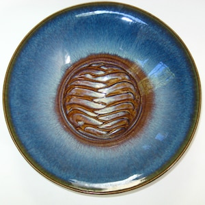 May include: A handmade ceramic bowl with a glossy blue glaze. The center features a textured, circular design in shades of brown and cream. The rim has a dark brown edge, and the bowl is round.