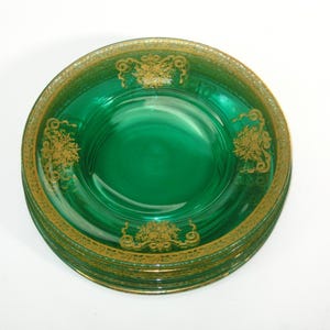 May include: A stack of emerald green glass plates with gold filigree detailing. The plates have a deep, rich color and a decorative gold border. The gold design includes floral and scroll motifs, adding a touch of elegance to the tableware.