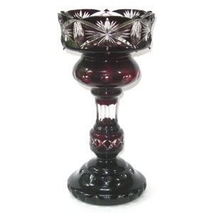 May include: A tall, dark red glass vase with a cut crystal design. The vase has a wide, round base and a narrow neck.