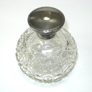 May include: A clear, round glass perfume bottle with a silver-colored lid. The bottle has a textured, diamond-like pattern. The lid is circular with a textured design. The bottle is empty.