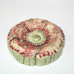 May include: A round ceramic flower frog with a pale green base and a textured surface featuring deep red, radiating brushstrokes. The top surface has a central circular section surrounded by eight evenly spaced holes for arranging stems.