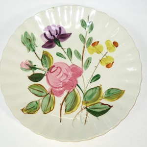 May include: Off-white ceramic plate with a scalloped edge, hand-painted with a floral design. The flowers include a pink rose, purple and yellow blooms, and green leaves. A decorative piece for serving or display.