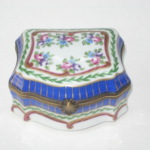 May include: A small, white porcelain trinket box with a blue and gold striped base. The lid is decorated with a floral design in shades of pink, blue, and green.