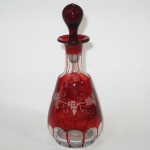 May include: A red glass decanter with a matching stopper. The decanter has a bulbous body with etched floral designs and vertical stripes. The stopper is a rounded shape. The decanter is likely for wine or spirits.