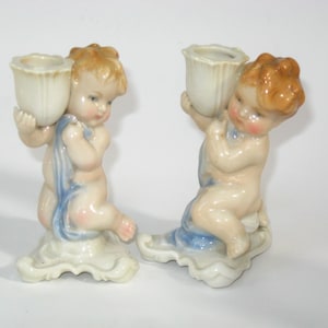 May include: A pair of vintage ceramic cherub candle holders. Each cherub is nude with peach skin, holding a white tulip-shaped candle cup. They have light brown hair and are draped with a blue cloth. The bases are white and cloud-like.
