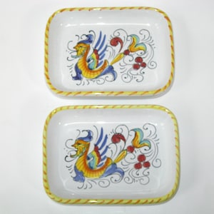May include: Two rectangular ceramic dishes with rounded corners, featuring a white base and a yellow rope-patterned rim. Each dish displays a hand-painted, colorful mythical creature design in blue, yellow, and red, accented by black swirls.