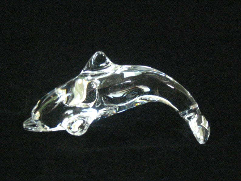 Op de afbeelding: Een helder glazen dolfijnbeeldje met een gebogen lichaam en een rugvin. De dolfijn is afgebeeld in een zwempositie, met zijn staart omhoog gebogen. Het beeldje is gemaakt van transparant glas.
