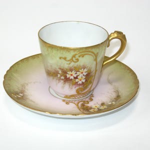 May include: A vintage teacup and saucer set. The cup and saucer have a pink and green gradient with gold trim. The cup features a floral design with white flowers and brown centers. The saucer has a scalloped edge.
