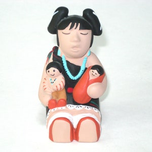 May include: A ceramic figurine of a woman holding two children. The woman wears a black top, a turquoise necklace, and has black hair. One child is wrapped in a red blanket, and the other is in a tan outfit. The figurine has a painted design.