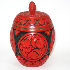 May include: A red and black decorative jar with a lid. The jar features intricate floral carvings in red against a black background. The lid has a small, rounded finial. The jar is likely made of a hard material.