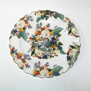 May include: White ceramic serving plate with a scalloped edge and gold trim. The plate is decorated with a floral pattern featuring white roses, orange fruit, and blue and green leaves. The design is repeated around the plate.