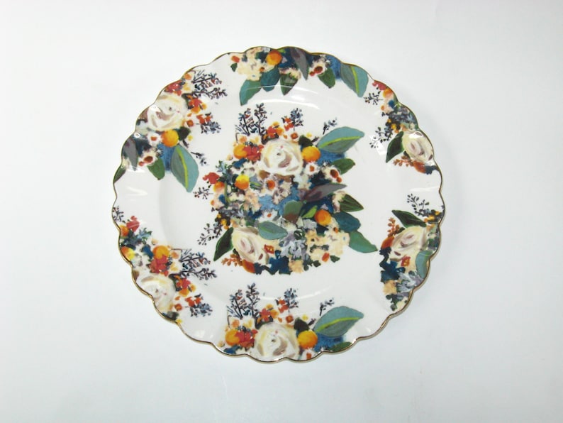 May include: White ceramic serving plate with a scalloped edge and gold trim. The plate is decorated with a floral pattern featuring white roses, orange fruit, and blue and green leaves. The design is repeated around the plate.