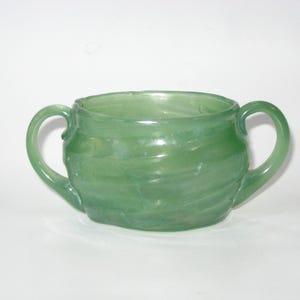 May include: A green glass sugar bowl with two curved handles. The bowl has a rounded shape and a textured, swirled design. The glass has a translucent quality, allowing light to pass through. The sugar bowl is set against a white background.