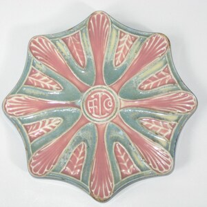 May include: A star-shaped ceramic dish with a pink and blue glaze. The dish has a raised design of leaves and a central circle with the letters "F.L.C." in white.