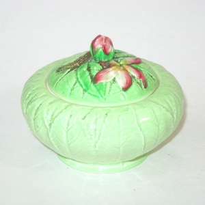 May include: A light green ceramic container with a lid. The lid is decorated with a pink and red flower and a green leaf design. The container has a leaf pattern and a round base.