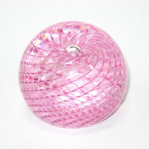 May include: A spherical glass paperweight featuring a translucent pink hue with an intricate, swirling white lattice pattern embedded throughout the surface. The object has a smooth, polished finish and a small, clear glass bubble at the center top.