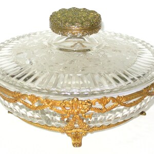 May include: Clear glass vanity jar with a ribbed design and a decorative gold-colored metal rim and accents. The lid is topped with a small, round, gold-colored metal piece. The jar sits on three small feet.
