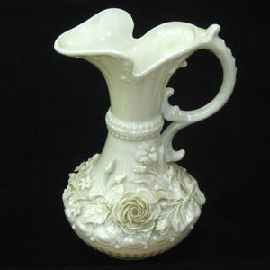 May include: An off-white ceramic pitcher featuring a glossy, iridescent finish. The body is adorned with raised, sculpted floral details including roses and leaves. It has a flared, scalloped rim and a curved handle with decorative accents.