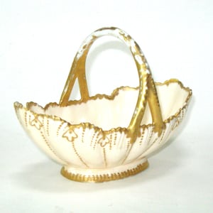 May include: Cream-colored ceramic basket with a scalloped rim and a gold handle. The basket is adorned with gold floral accents and a gold base. A decorative piece for display or serving.