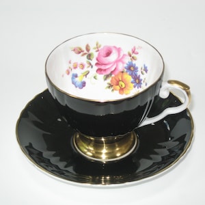 May include: A black teacup and saucer set with a white floral design. The cup has a gold rim and a gold base. The saucer is black with a gold rim.