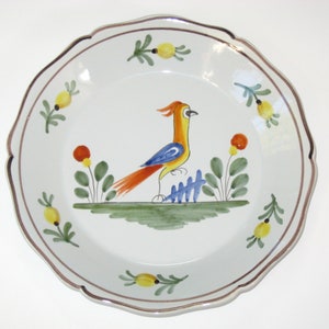 May include: A round ceramic plate with a scalloped edge and a brown rim. The center features a hand-painted yellow, blue, and orange bird standing on a green patch of grass. The border is decorated with small yellow fruit and green leaves.