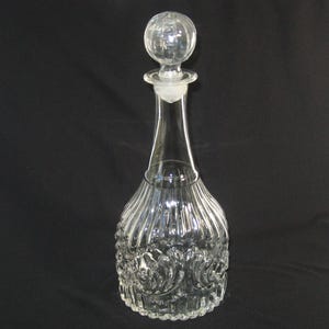 May include: A clear glass decanter featuring a spherical stopper and a long, slender neck. The base of the decanter is wide and rounded, decorated with vertical fluting and embossed scrollwork patterns that catch the light.