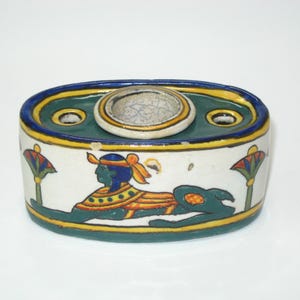 May include: An oval-shaped ceramic ashtray with an Egyptian theme. The ashtray features a central circular depression and two smaller holes. The exterior is decorated with a sphinx and lotus flower motifs, with blue, yellow, green, and orange accents.