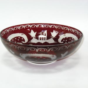 May include: A ruby red glass bowl with white etched designs. The bowl features a repeating pattern of swirls and stylized birds. The interior has a central design of a building and birds. A decorative piece for serving or display.