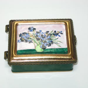 May include: A rectangular metal trinket box with a hinged lid featuring an enameled painting of blue irises in a green vase against a light pink background. The name Vincent is inscribed on the bottom right. The box has a dark green base.