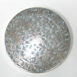May include: Circular silver-toned decorative plate with an intricate floral and vine design. The plate has a light blue background and is made of metal. The design is symmetrical and detailed, suitable for home decor.