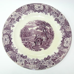 May include: A round, antique-style plate with a purple and white floral border. The center features a purple landscape scene with a building, trees, and a church. The plate has a cream-colored center and a decorative edge.