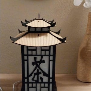 May include: A wooden lamp shaped like a pagoda with a black frame and a white translucent panel with the Chinese character for "tea" on it. The lamp is sitting on a wooden surface.