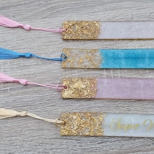 Op de afbeelding: Vier handgemaakte bladwijzers met bladgoudaccenten en gekleurd hars. De bladwijzers hebben kwastjes in roze, blauw, wit en roze. Eén bladwijzer heeft de tekst "Super Momma" erop.