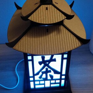 May include: A wooden lantern with a blue light shining through the intricate design. The lantern has a traditional Chinese roof and the word "茶" (tea) is visible on the front.