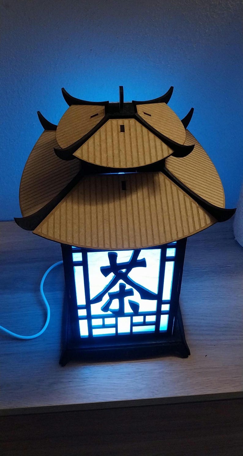 May include: A wooden lantern with a blue light shining through the intricate design. The lantern has a traditional Chinese roof and the word "茶" (tea) is visible on the front.