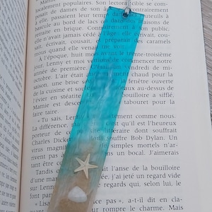 Op de afbeelding: Een turquoise blauwe en witte harsboekmerk met een ster en schelp ingebed in de hars. Het boekmerk is rechthoekig en heeft een kwast aan de bovenkant.