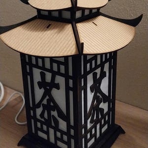 May include: A wooden lamp with a black frame and a white paper insert. The lamp has a pagoda-style roof and the word "茶" is printed on the paper insert.