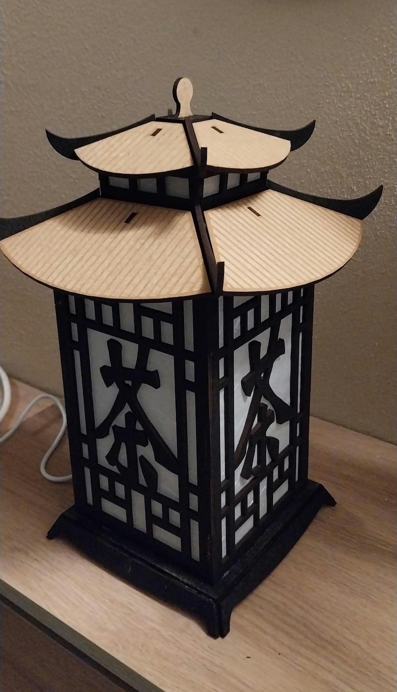 May include: A wooden lamp with a black frame and a white paper insert. The lamp has a pagoda-style roof and the word "茶" is printed on the paper insert.
