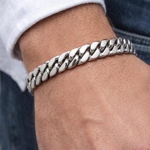 May include: A silver curb chain bracelet. The bracelet is made of interlocking links and is worn on a wrist. The bracelet is a bright, shiny silver color and appears to be made of metal.