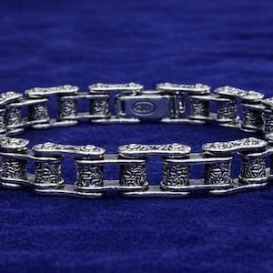 May include: Silver-toned metal bracelet with a chain link design. The links are detailed with ornate patterns and connect with a clasp. The bracelet is displayed on a dark blue velvet surface.