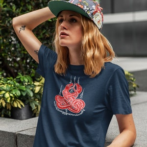 Peut inclure: Un t-shirt bleu marine avec un motif blanc représentant un serpent rouge avec des caractères thaïlandais. Le serpent est enroulé en cercle, la tête tournée vers le haut. L'écriture est en caractères thaïlandais traditionnels.